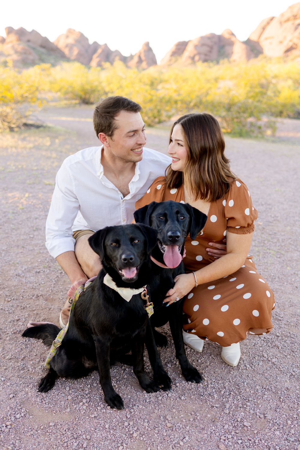 Happy couple with their two black Labrador dogs sitting outdoors against red desert mountains during sunset.