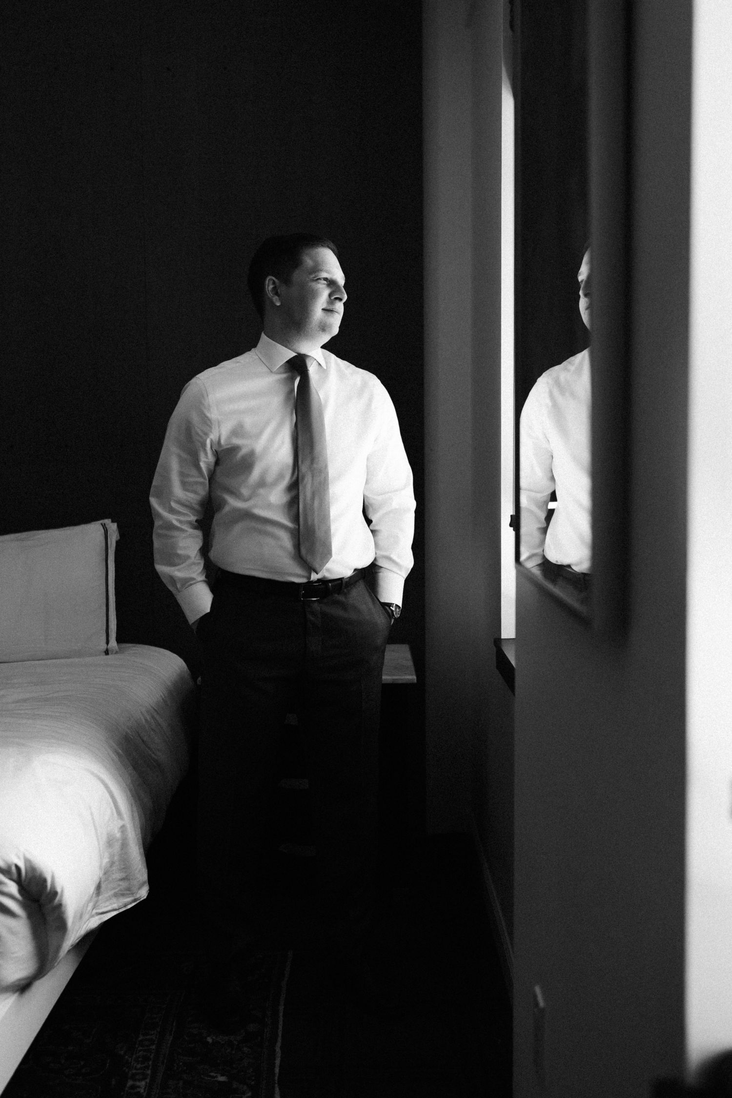 Black and white portrait of person in white dress shirt and tie looking pensively out window in moody lighting.