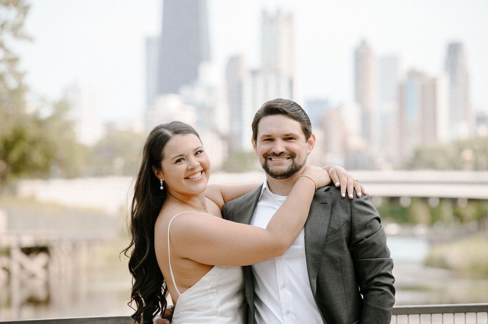 A couple in wedding attire poses together on a scenic overlook with a city skyline in the background.