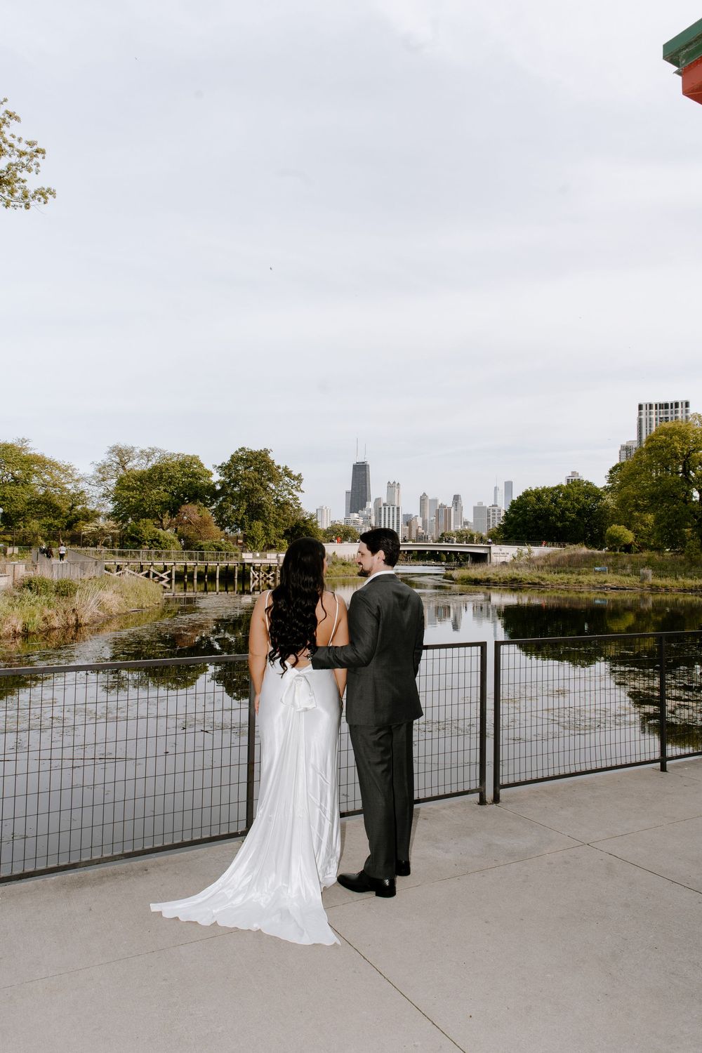 A couple shares a romantic moment on a waterfront promenade with a cityscape visible in the distance.