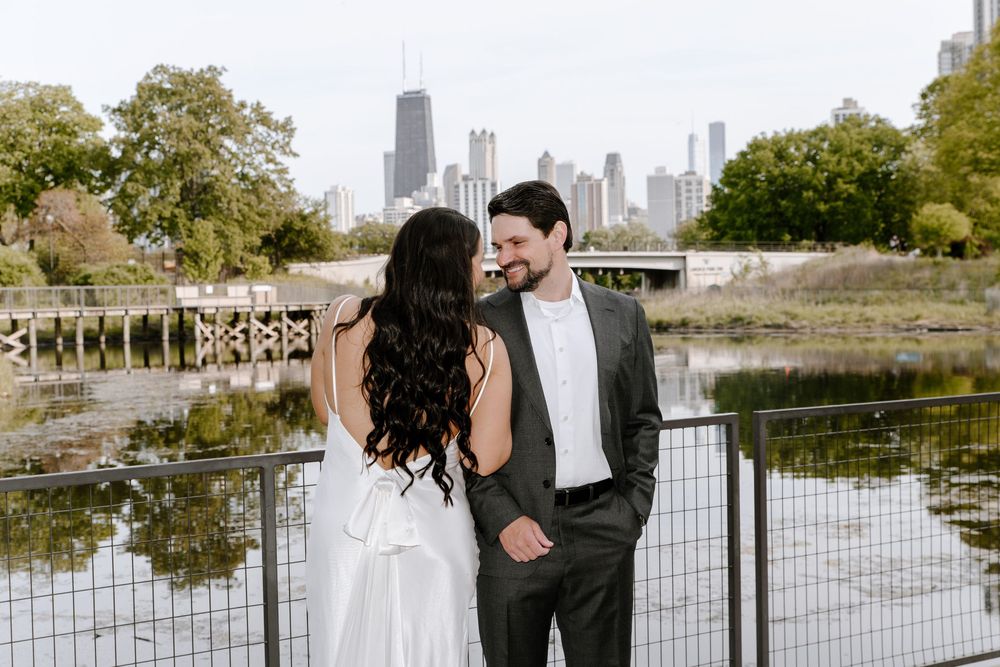 A couple in formal wedding attire embraces on a paved pathway with railings and an urban skyline behind them.
