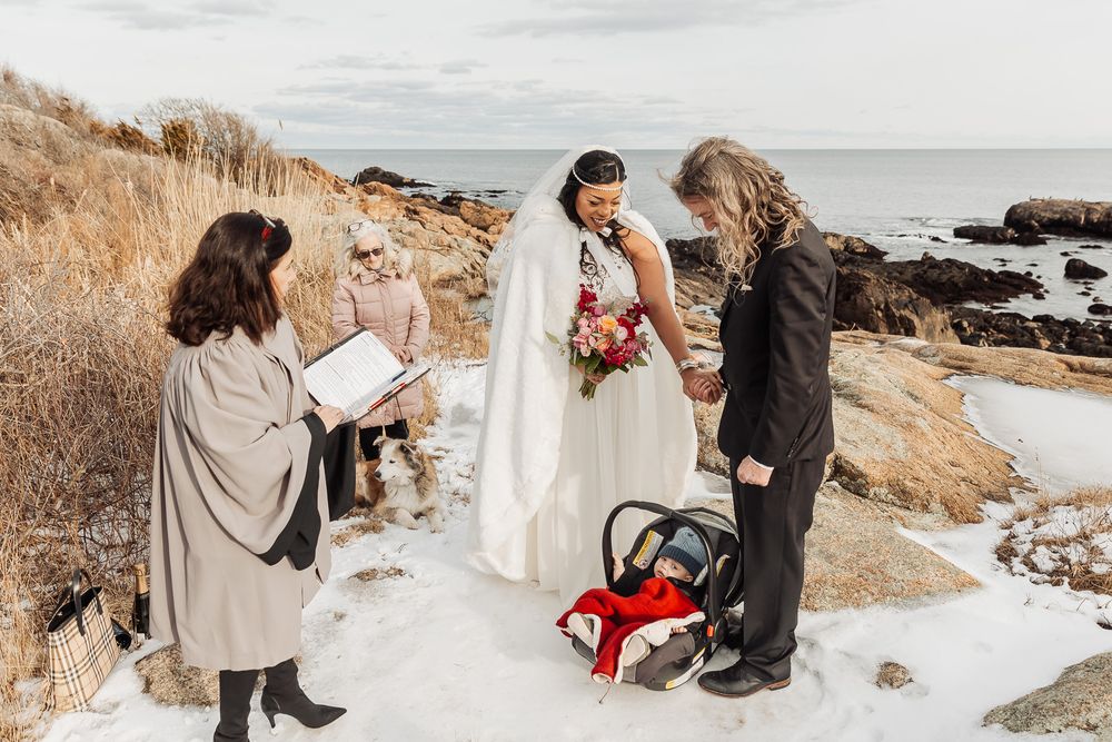 A small winter wedding ceremony takes place on a snowy rocky coastline with waves and dried grass in the background.