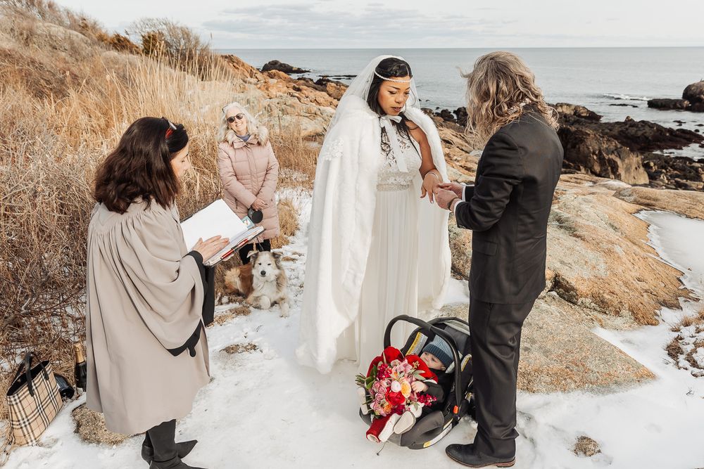 A winter wedding ceremony takes place on rocky coastal cliffs with attendees wrapped in warm layers.