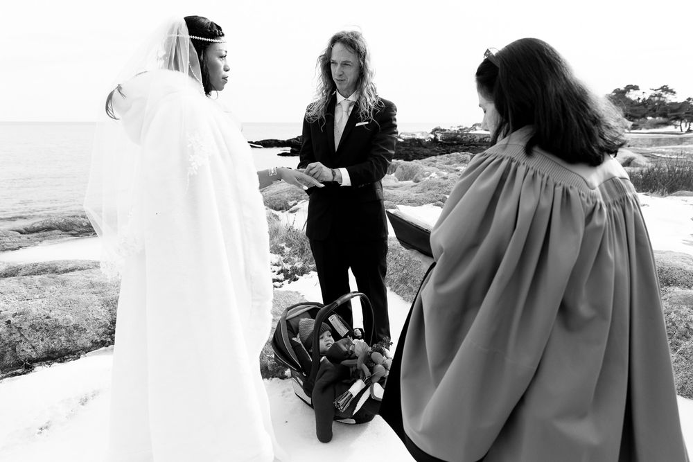 A black and white photograph of a couple exchanging vows during an intimate outdoor seaside wedding ceremony.