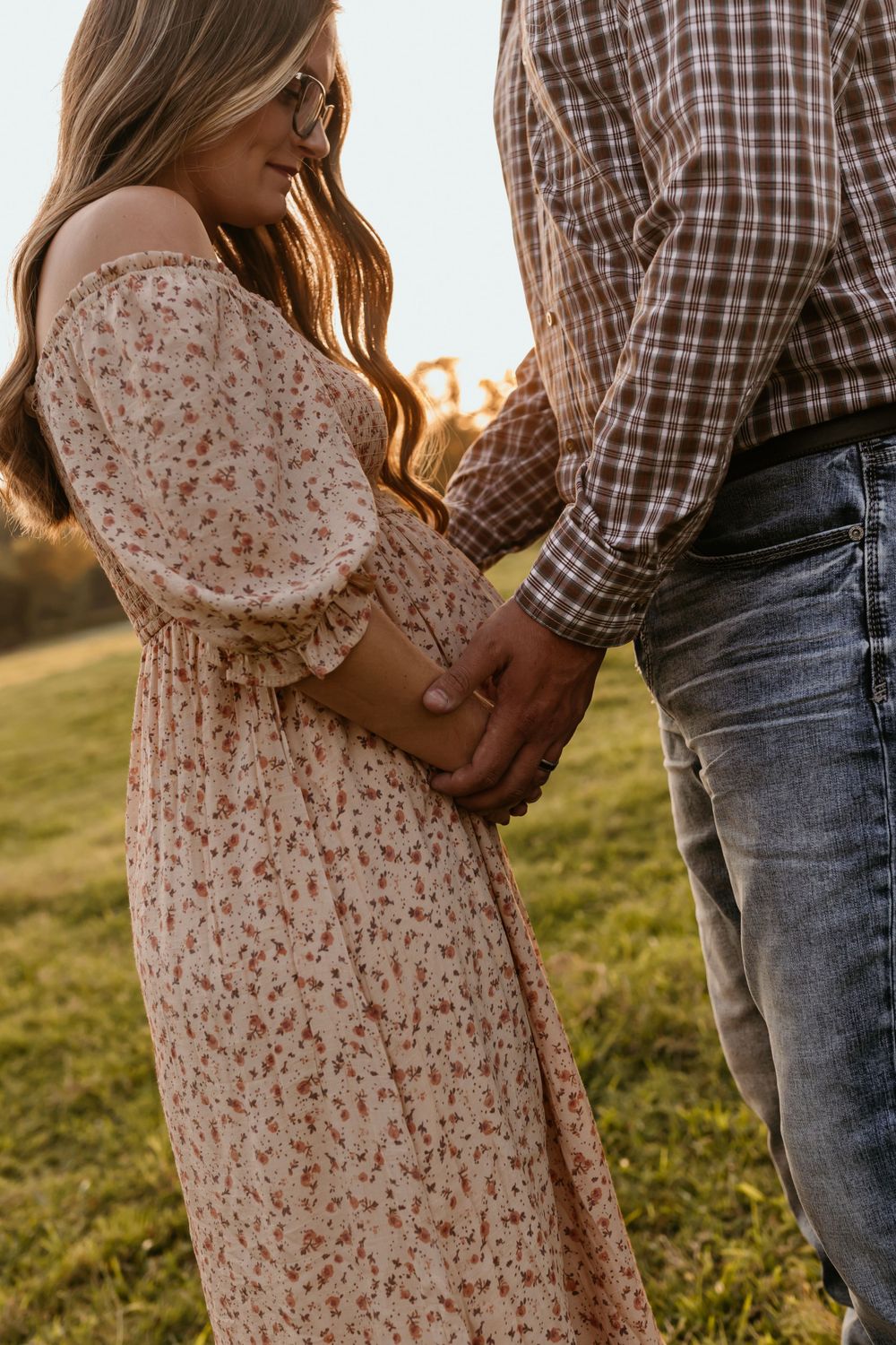 Couple holding hands in a sunlit field, wearing floral dress and plaid shirt.