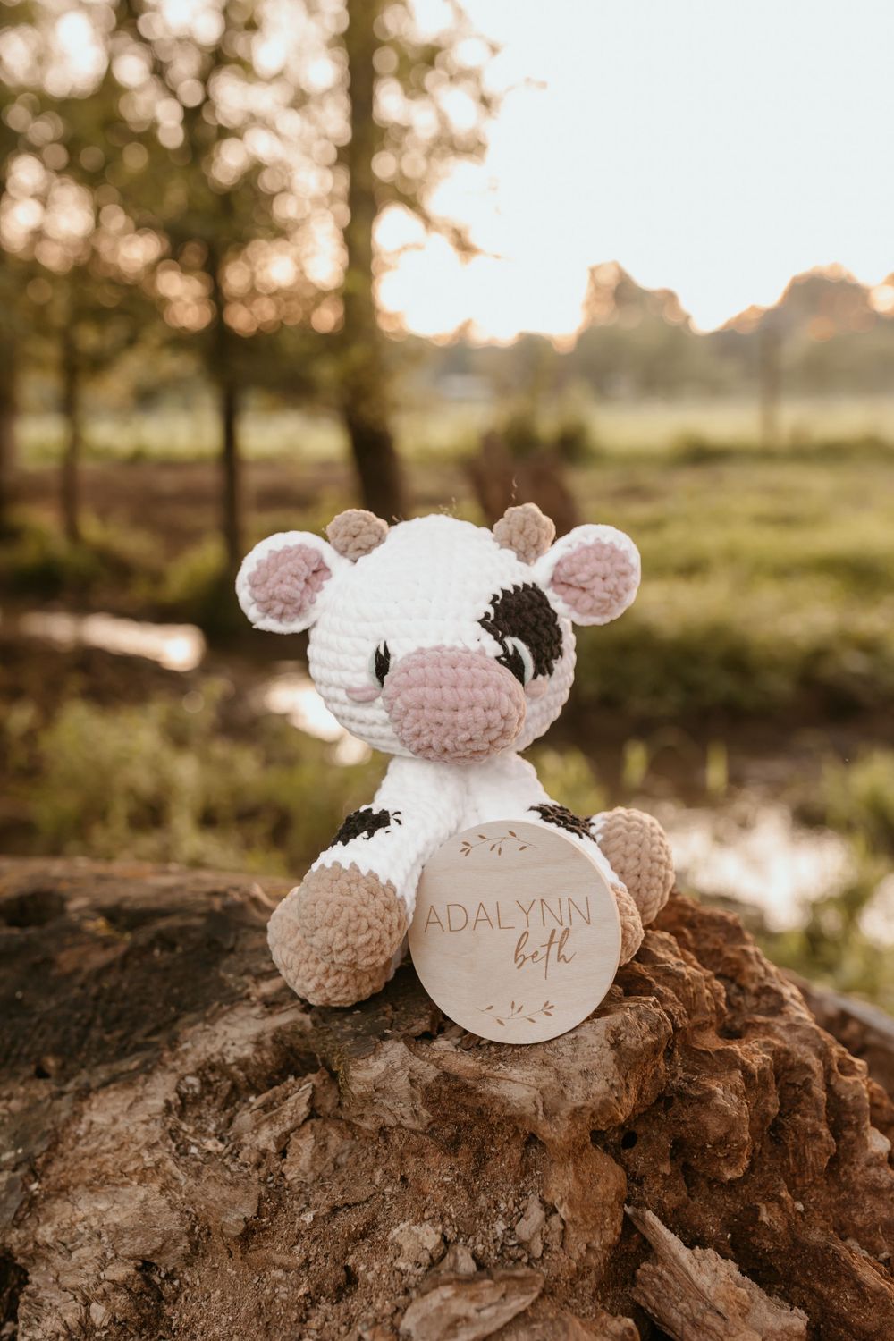 Cute crocheted white and pink cow plush toy sitting on a rock at sunset.