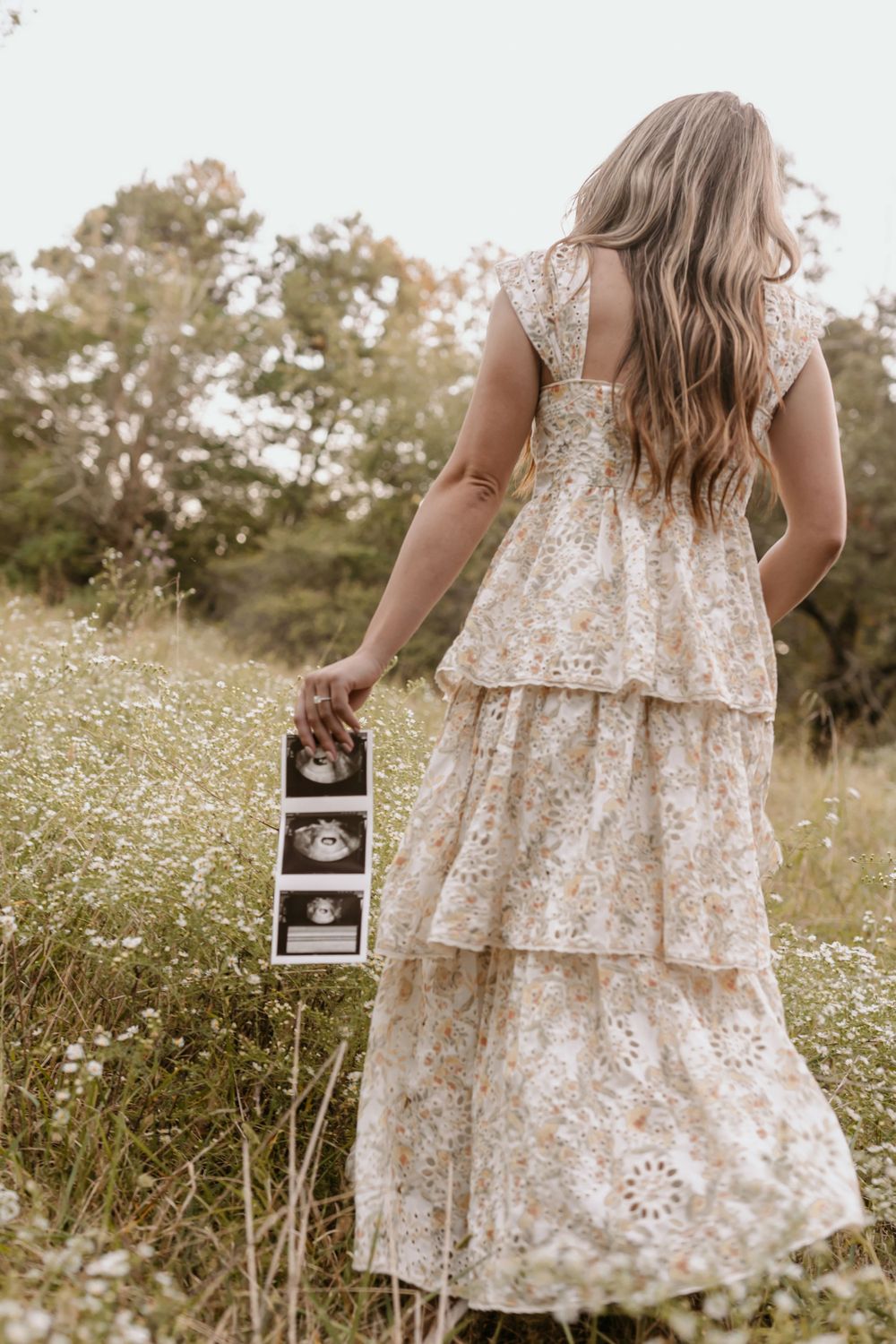 Pregnant woman in floral tiered dress holds ultrasound photos while standing in a wildflower field at sunset.