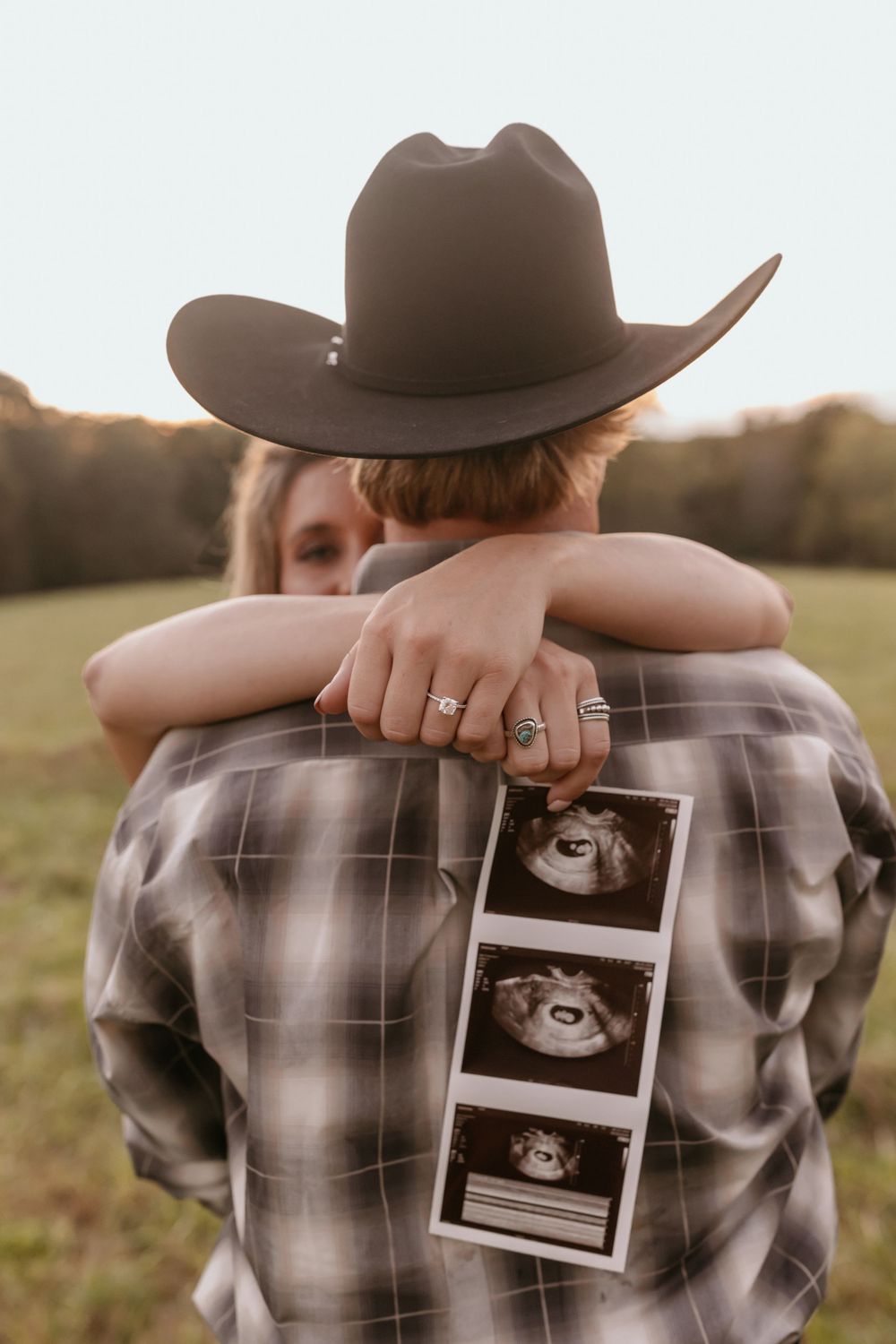 Person wearing cowboy hat holds ultrasound photos while embracing another in plaid shirt outdoors.