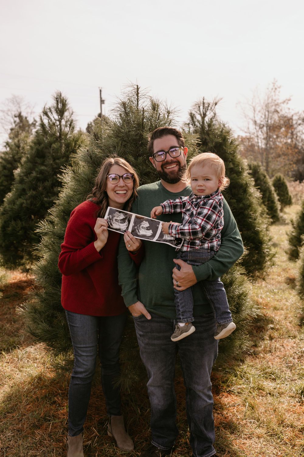 Family holding ultrasound photos while standing among Christmas trees at a tree farm.