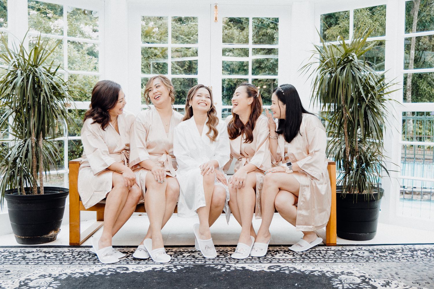 Five friends in matching silk robes and slippers sit together laughing captured by Apertured Media Auckland Wedding Photographer.