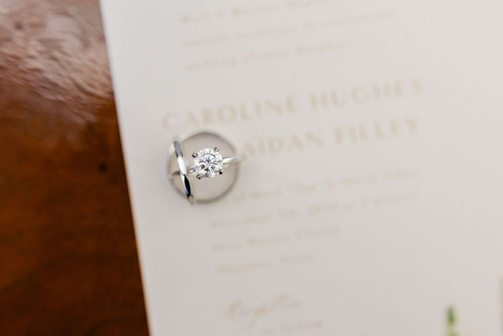 A sparkling diamond engagement ring photographed against a white invitation with elegant text.