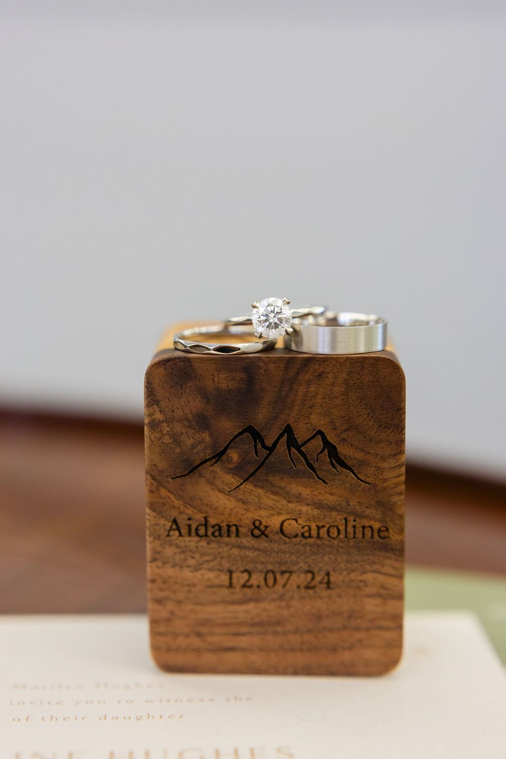 Wedding rings displayed on wooden box with mountain design and names engraved for December 2024 date.