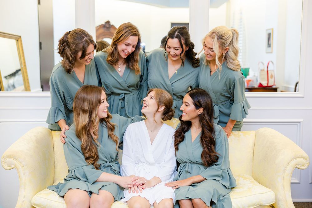 Bridesmaids in sage green robes gather around bride on yellow sofa during wedding preparations.