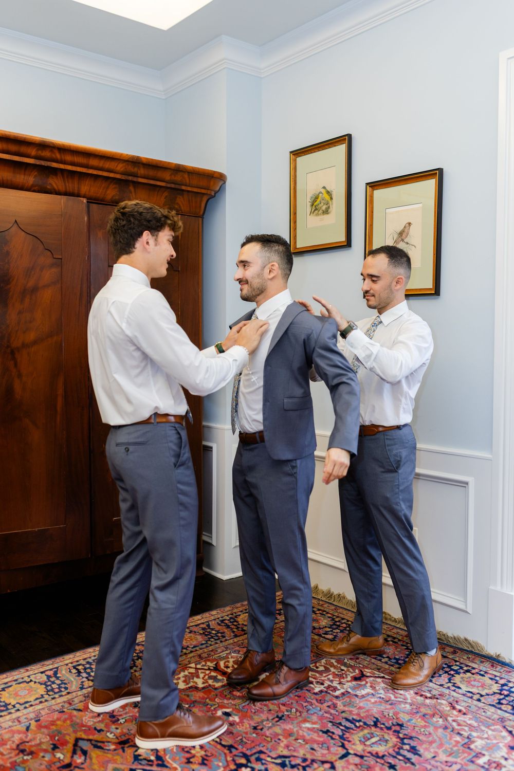 Groomsmen help adjust a suit and tie while getting ready in an elegant room with patterned carpet and wooden armoire.