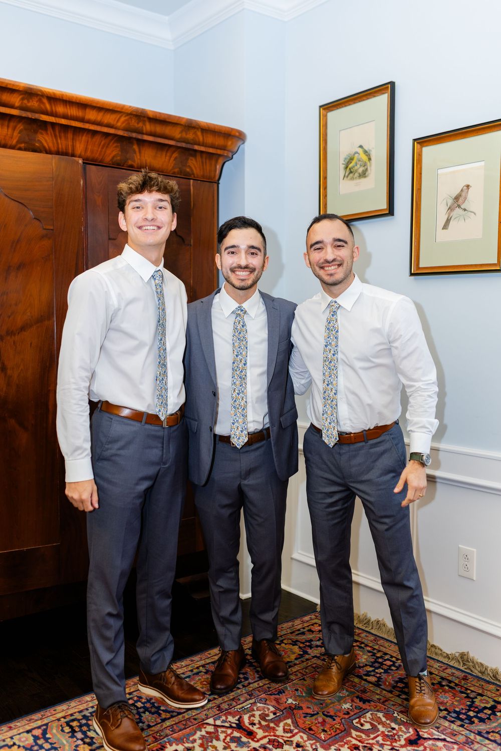 Three groomsmen in coordinating navy suits and patterned ties pose together in front of a wooden armoire.