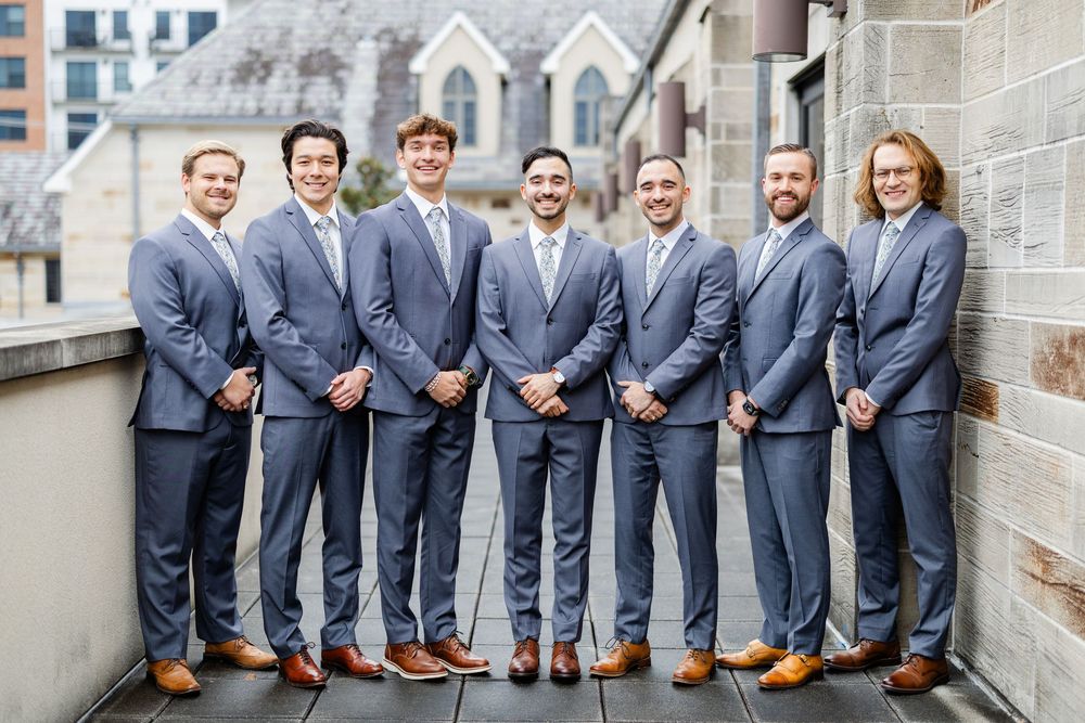 Seven groomsmen in matching navy suits and brown shoes stand together on an outdoor terrace.