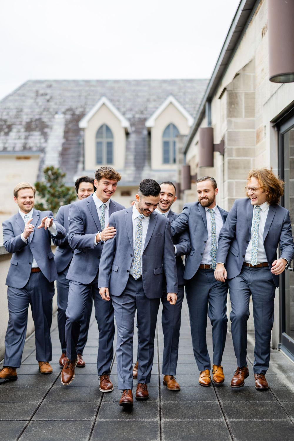 A group of groomsmen in gray suits and brown shoes share a candid moment walking together outside a church.
