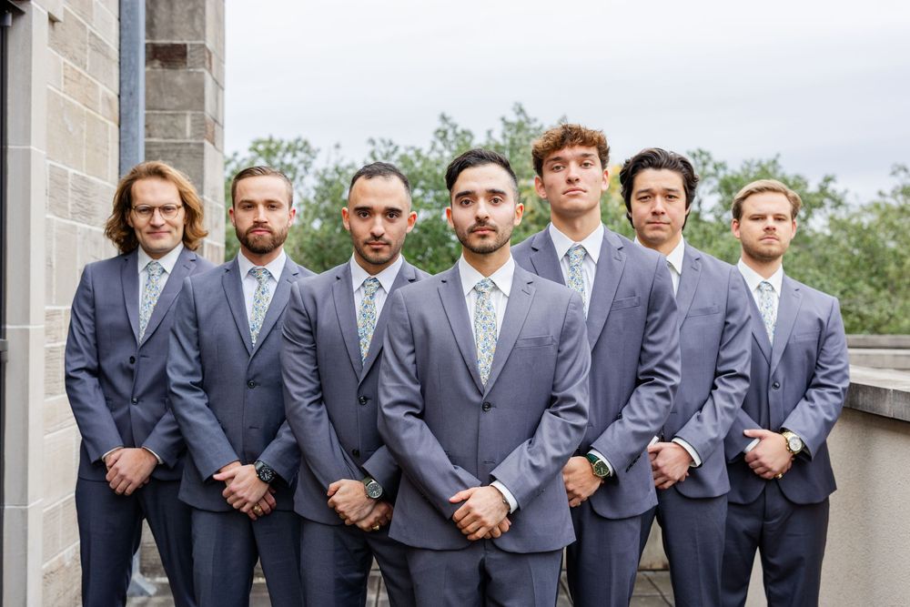 Seven groomsmen in matching navy suits pose together on an outdoor balcony.