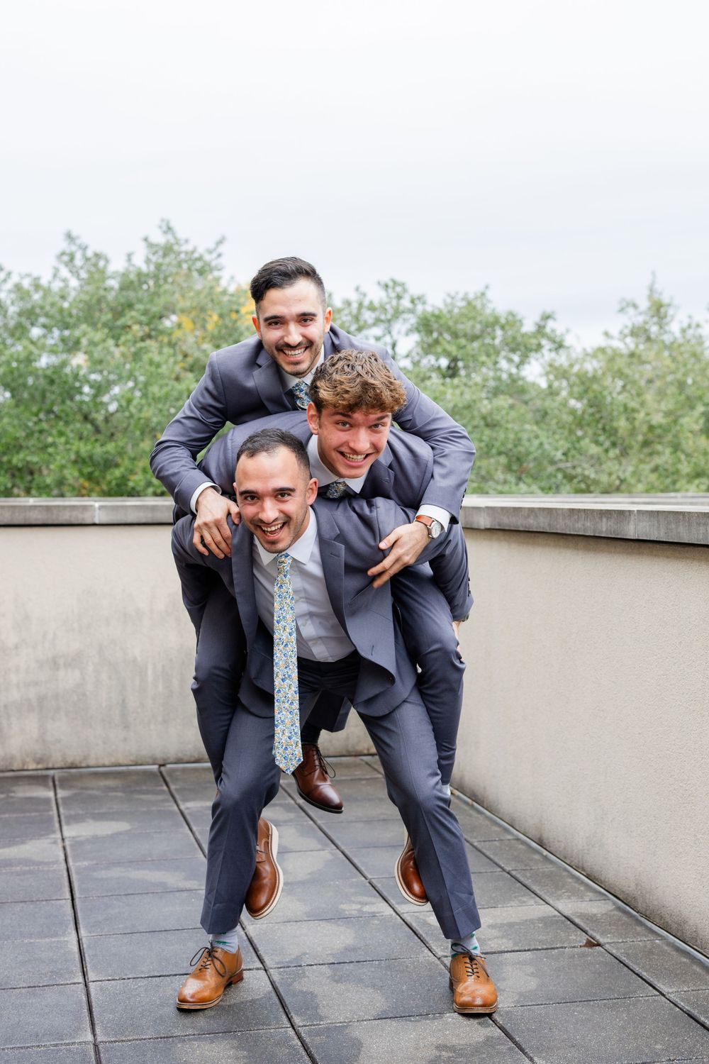 Three groomsmen in matching navy suits playfully pose together while piggybacking on a rooftop terrace.