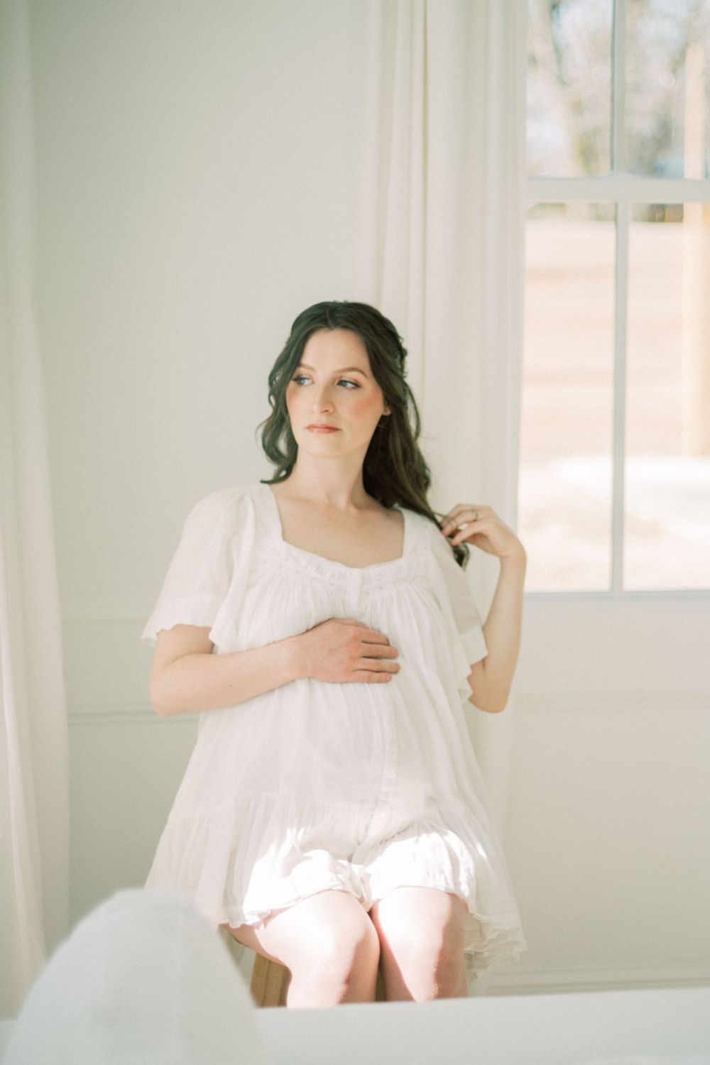 Woman in soft white dress sits near window in dreamy maternity photo with natural lighting.
