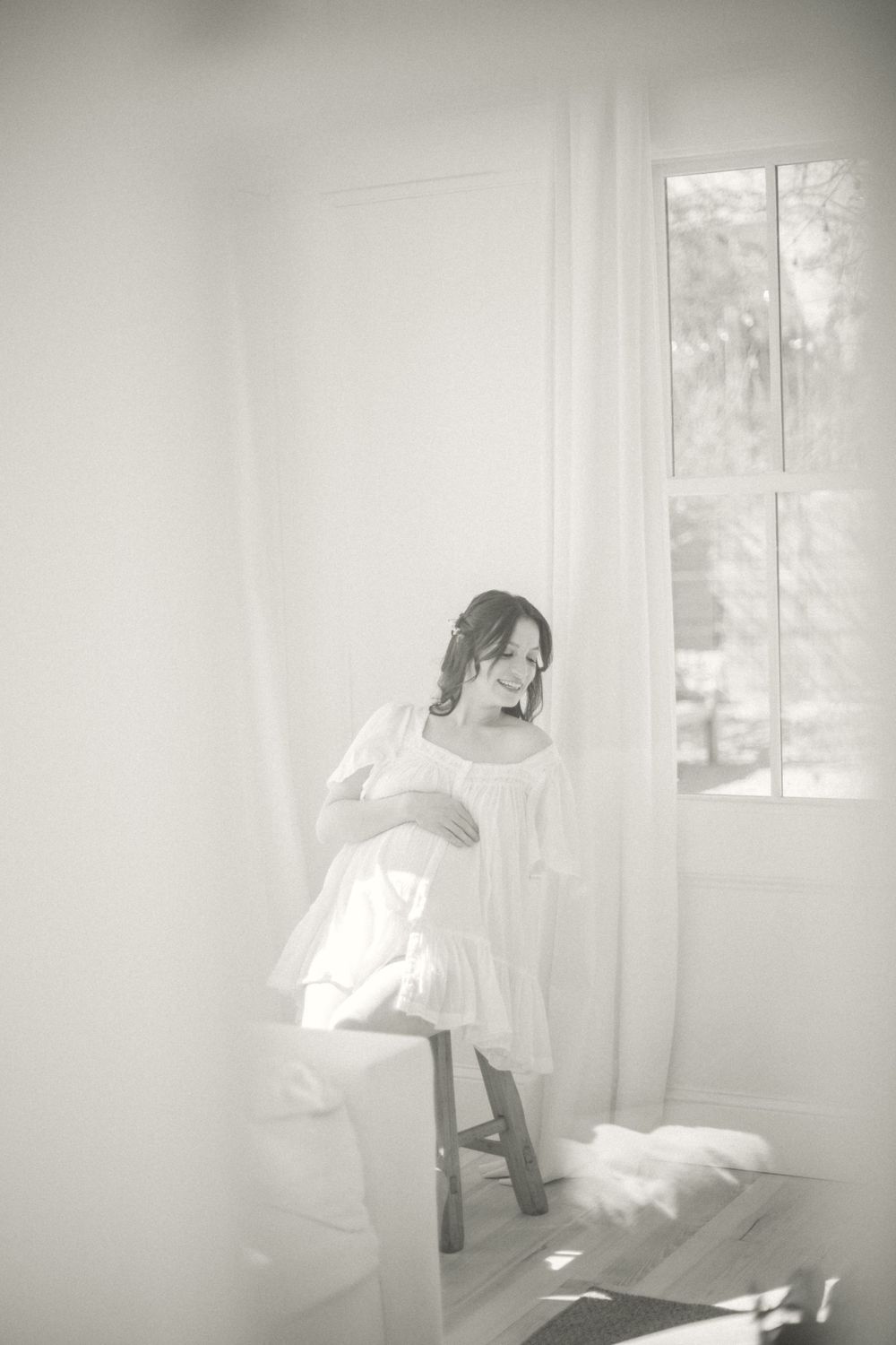 Dreamy black and white photograph of a person in a white dress sitting on a stool beside a sunlit window.