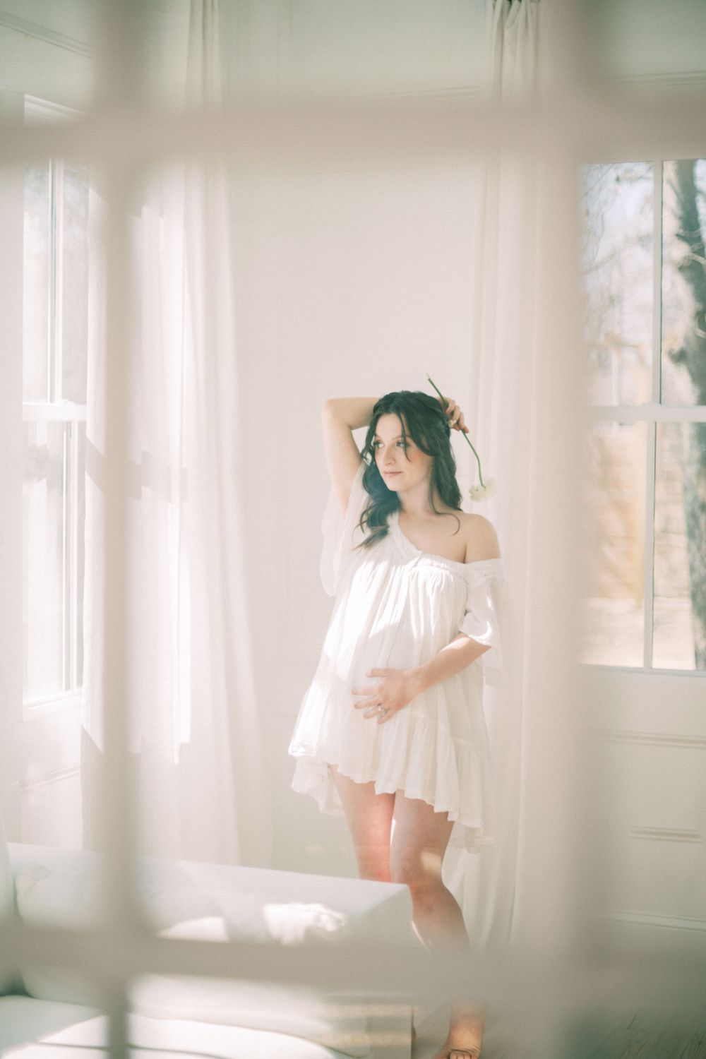 Soft light streams through white curtains onto a figure in a flowy off-shoulder white dress in a bright room.