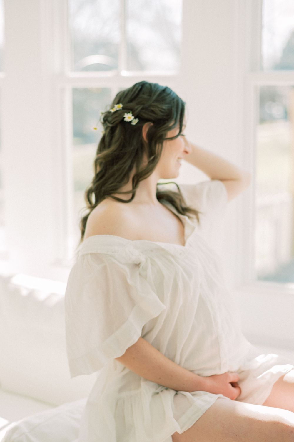 Ethereal bridal boudoir photo with flowing white dress and delicate flowers in soft window light.