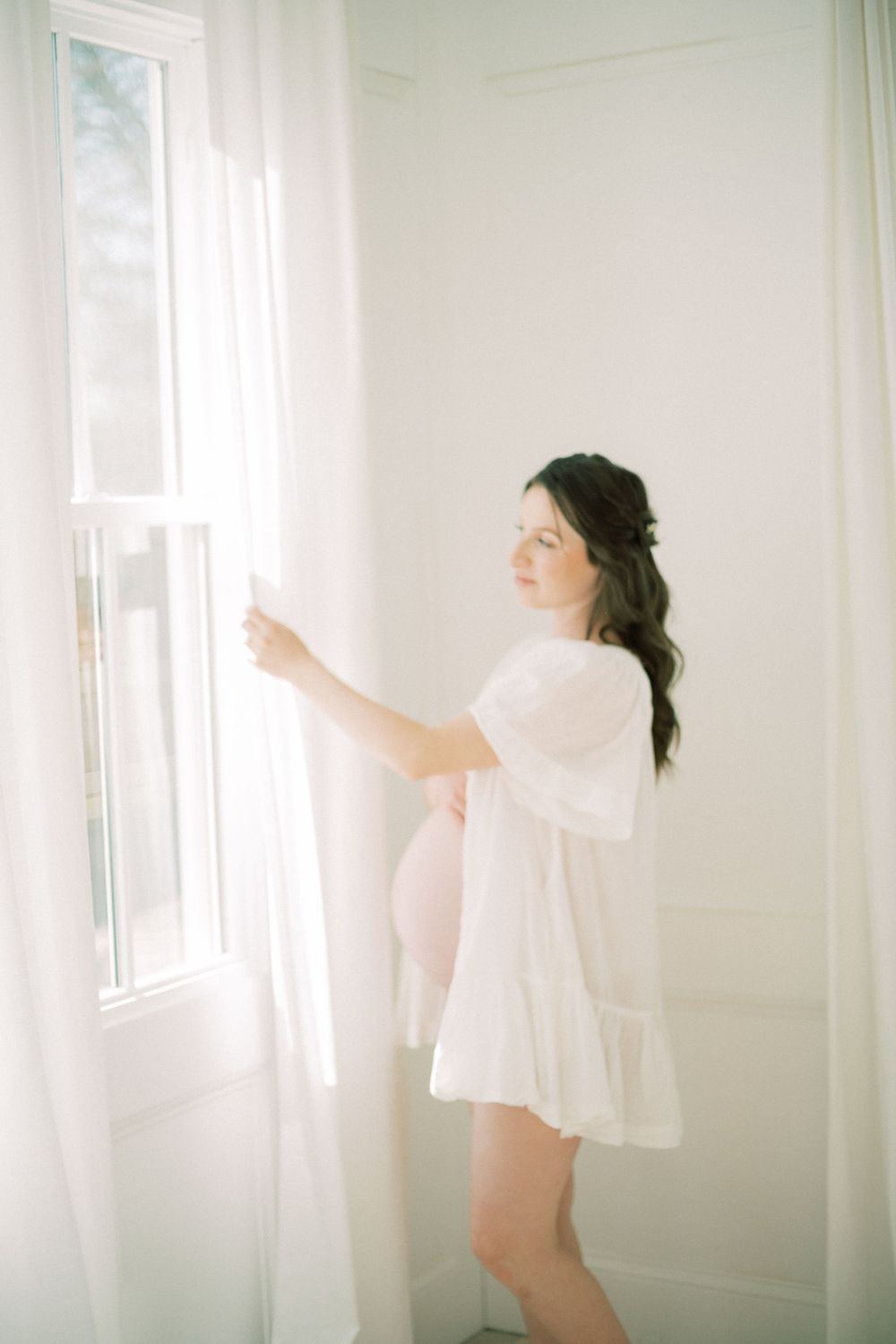 A woman in a flowy white dress stands by a bright window in a minimalist white room with ethereal lighting.