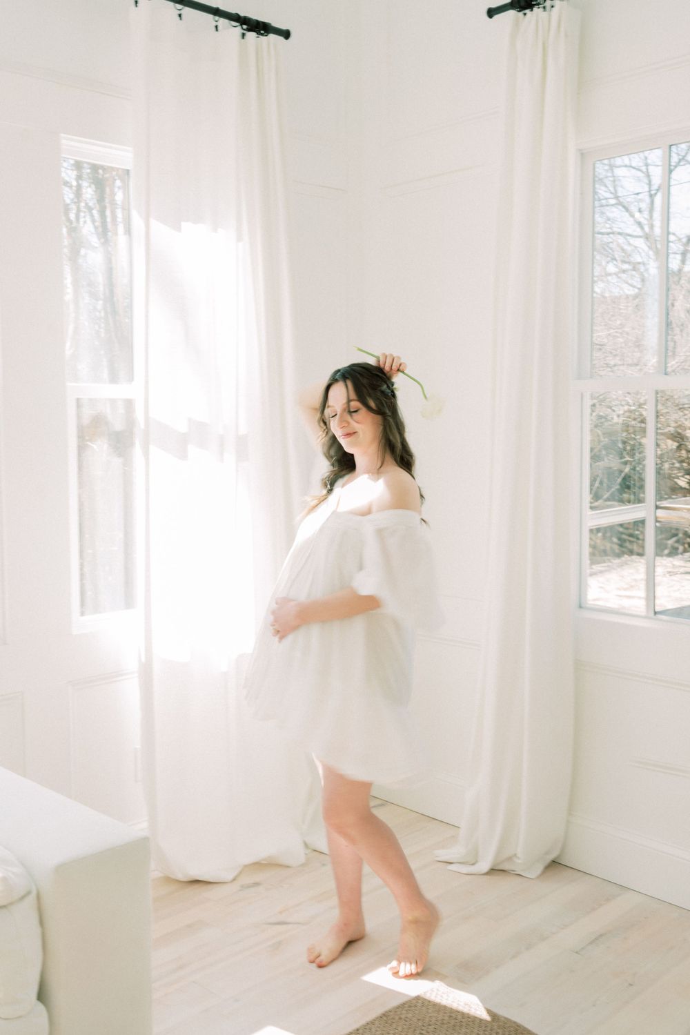 Ethereal figure in white flowing dress stands barefoot by sunlit windows with sheer curtains in bright minimalist room.