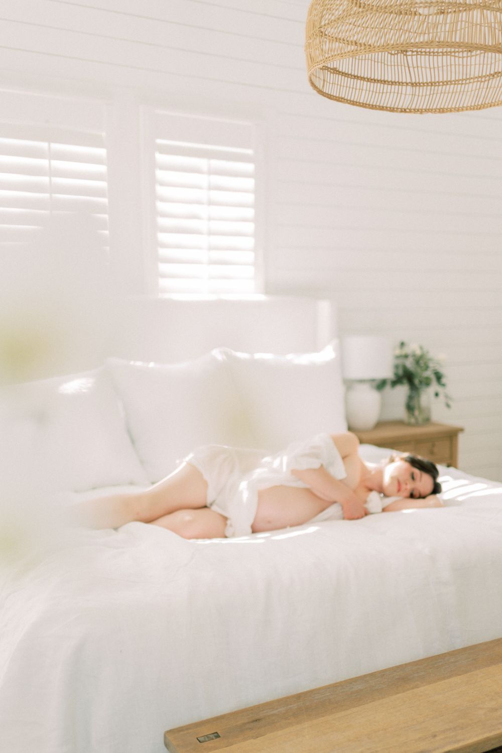 Serene bedroom scene with soft white bedding, natural light through shutters, and a rattan pendant lamp overhead.