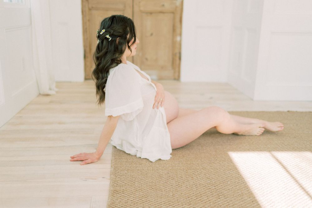 Woman in flowy white dress lounges on beige carpet in sunlit room with minimalist decor.