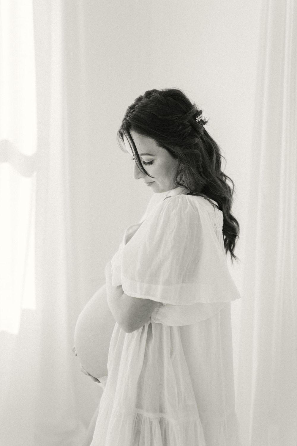 Black and white maternity portrait of an expectant mother in a flowing white dress standing by a window.