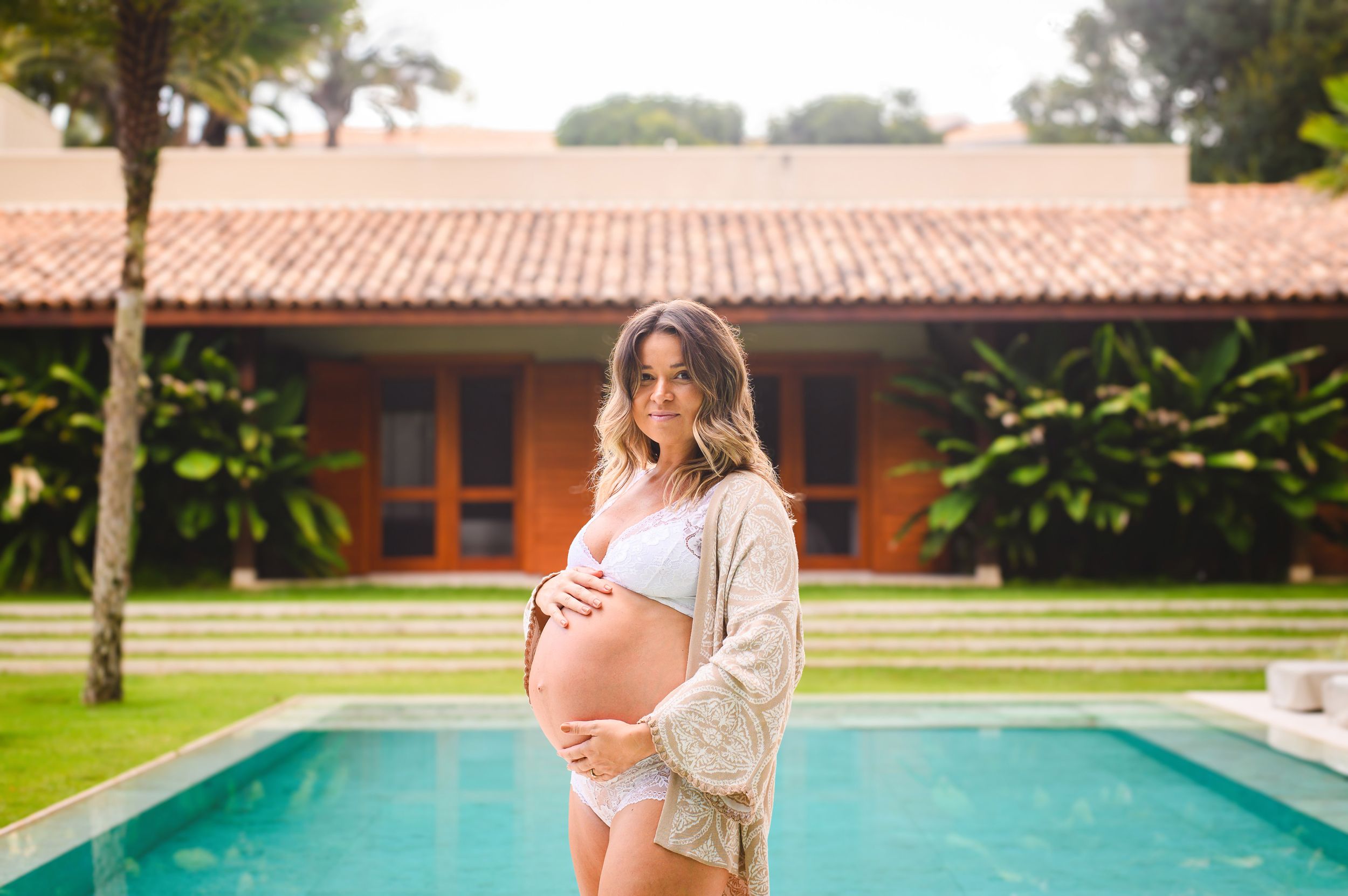 Pregnant woman in white lace robe stands beside swimming pool at tropical resort with terracotta roof building.