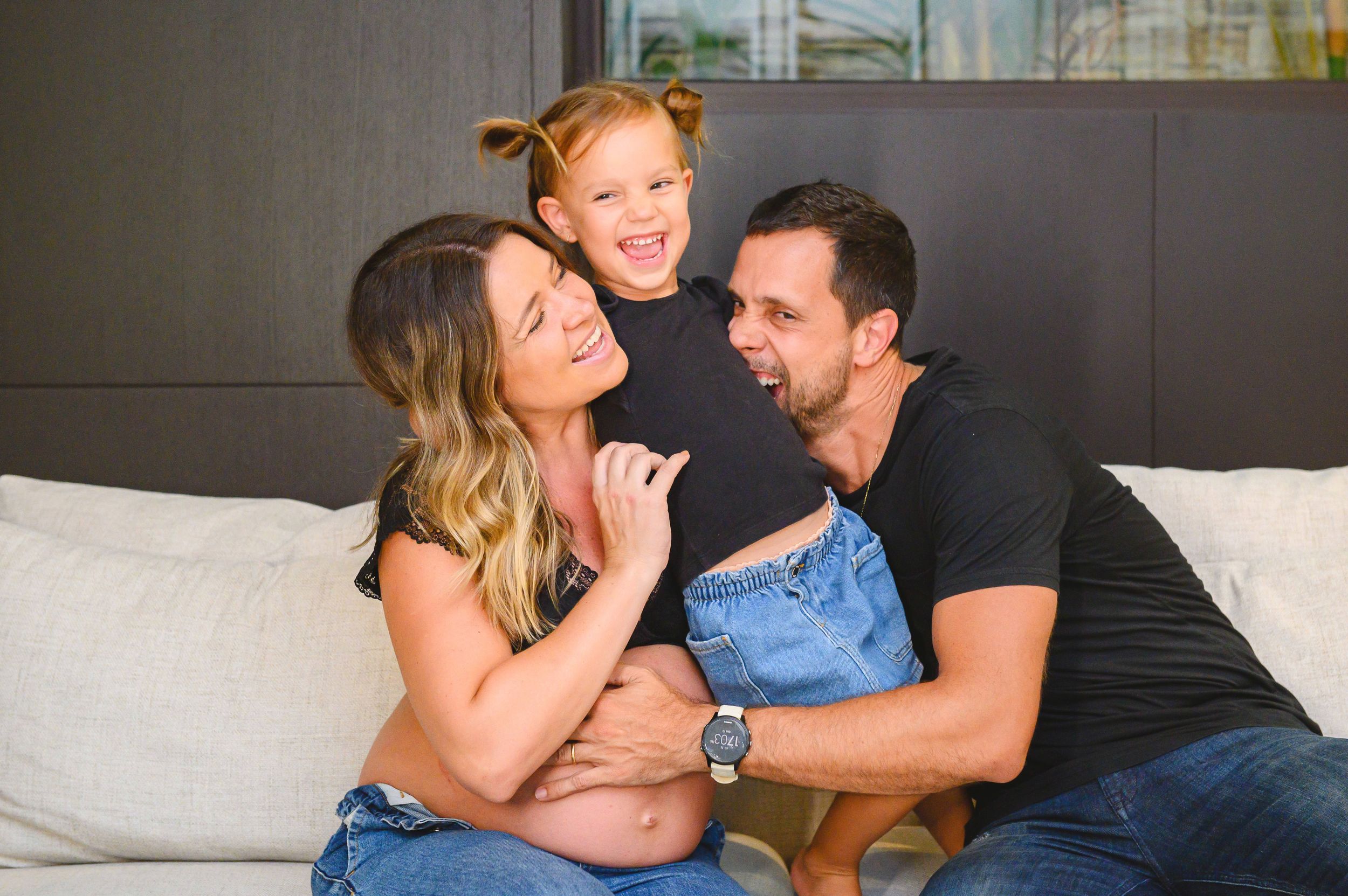 A happy family of three laughs together while sitting on a light gray couch in a modern living room.