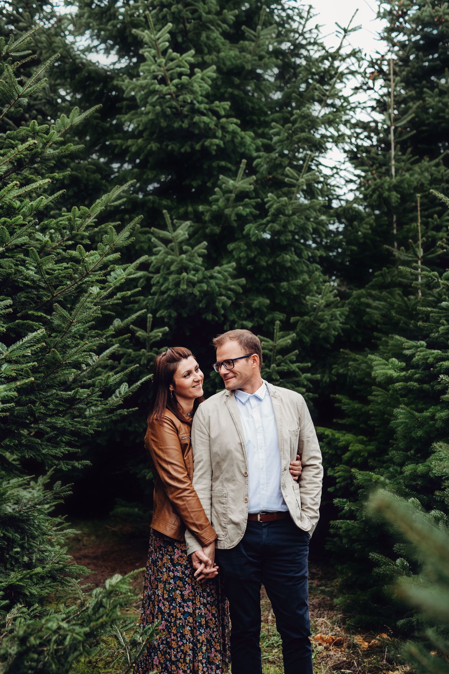 A couple shares an intimate moment among tall evergreen trees in a forest setting.