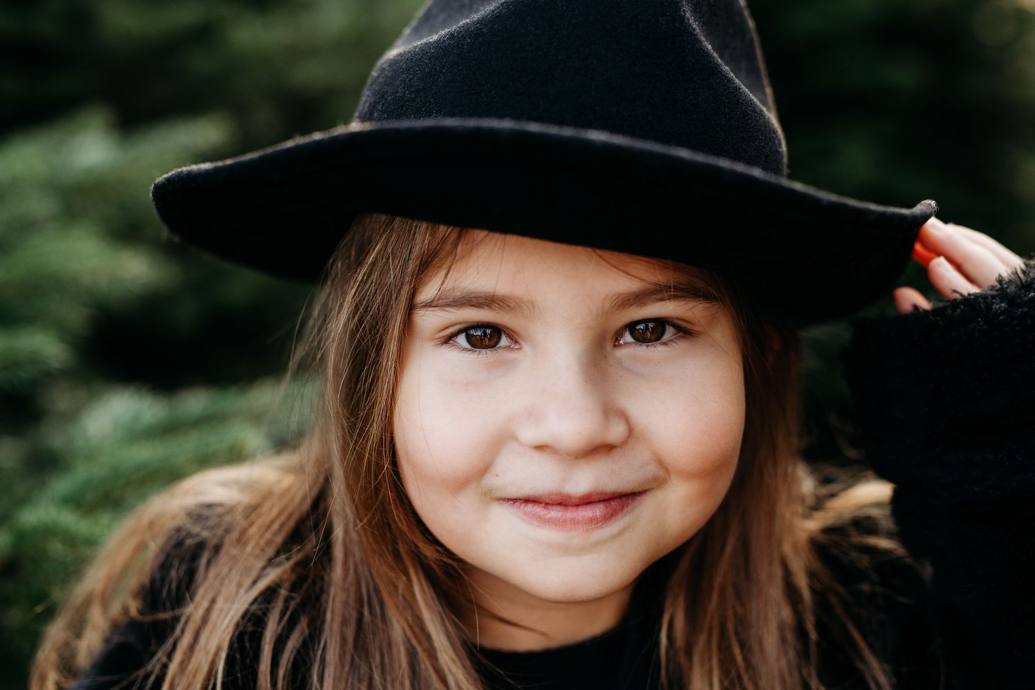 A young person wearing a black hat smiles warmly against an evergreen forest background.