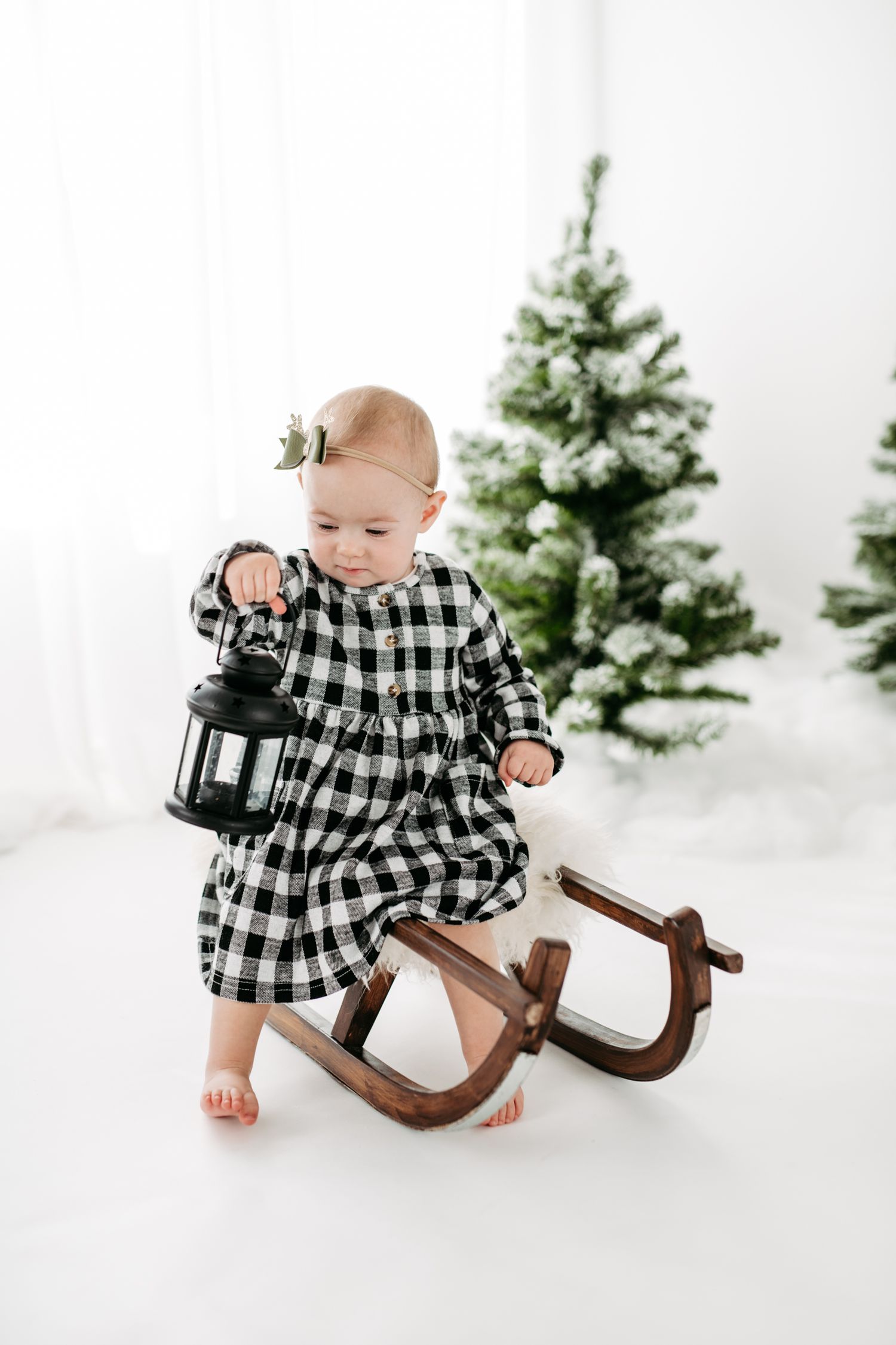 Small child in black and white checkered dress sits on wooden rocking horse with lantern near Christmas trees.