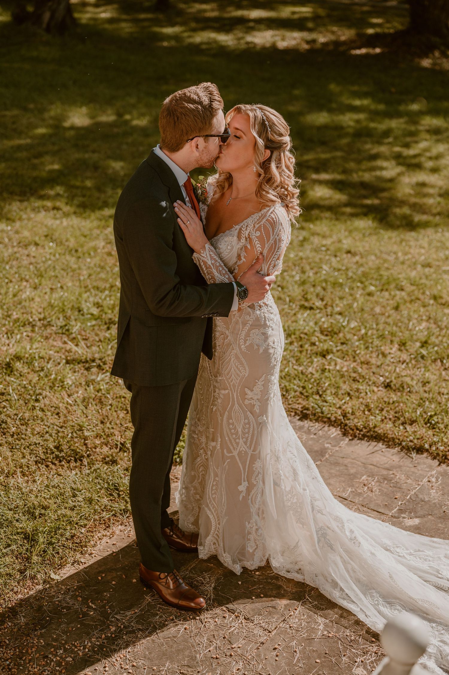Newlyweds share a tender kiss in a sunlit garden setting with the bride wearing an intricate lace gown with long train. by Samantha Campbell