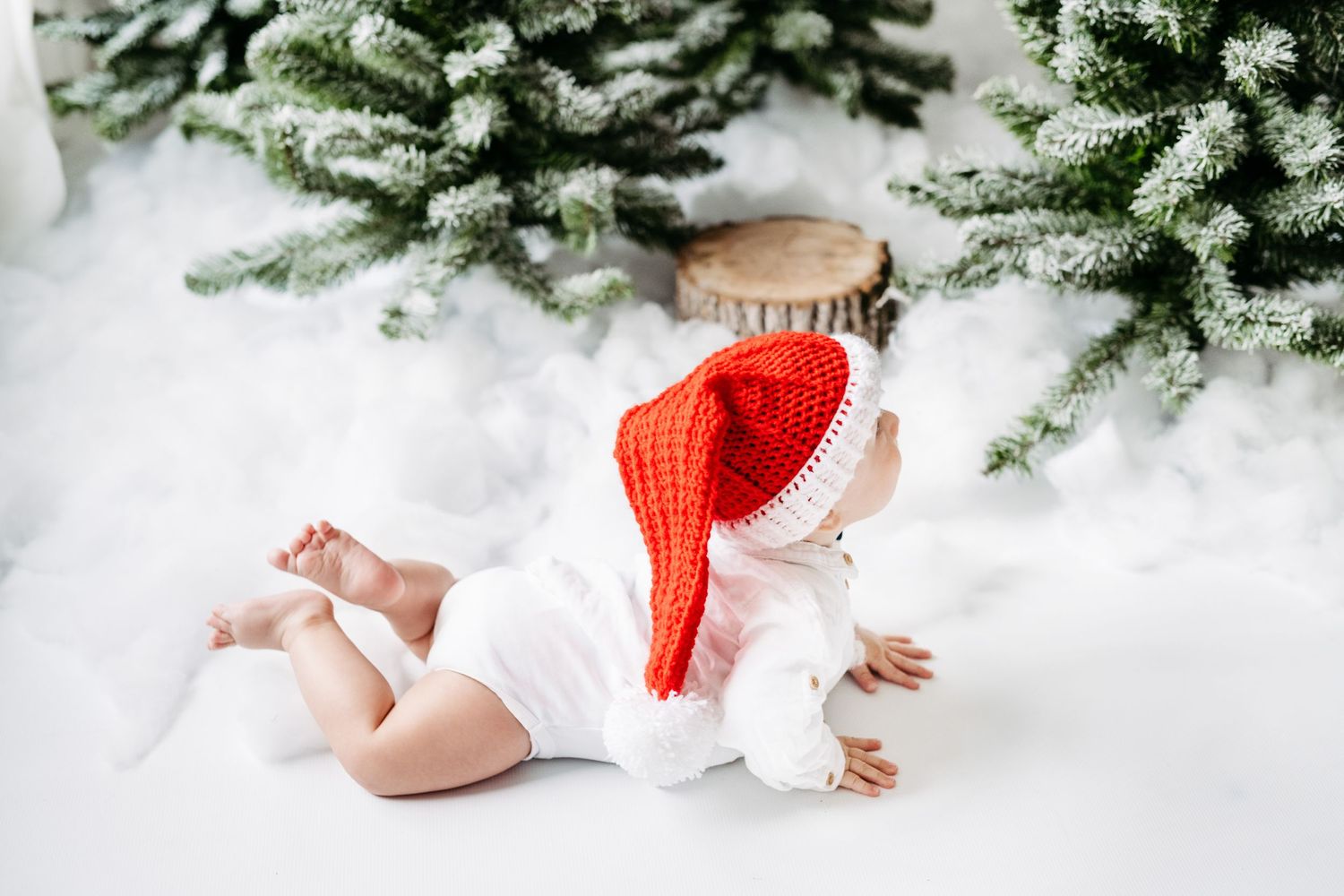 A baby in white clothing and red Santa hat lying on snowy ground near frosted Christmas trees.