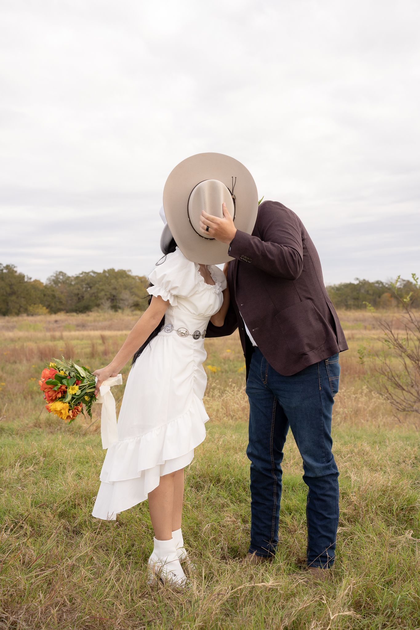 A couple in a countryside field share a romantic moment, with the bride holding sunflowers and wearing a flowy white dress.