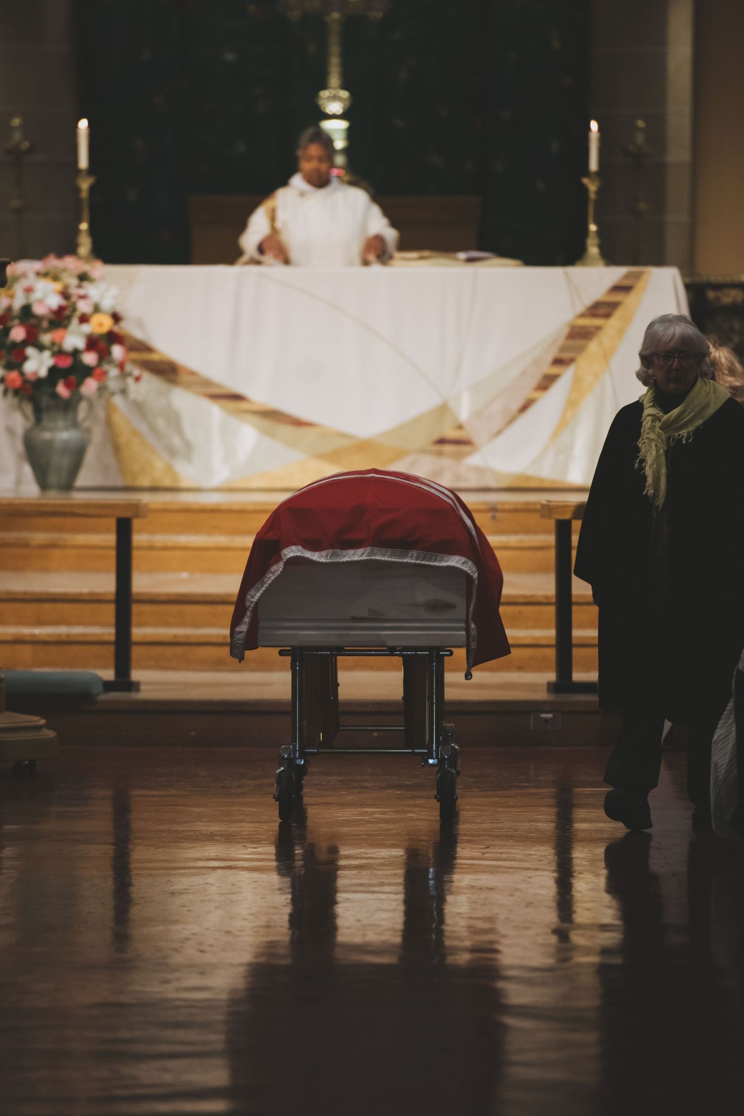 A church altar with a white cloth-covered table, candles, and flowers, with a draped casket in the foreground.
