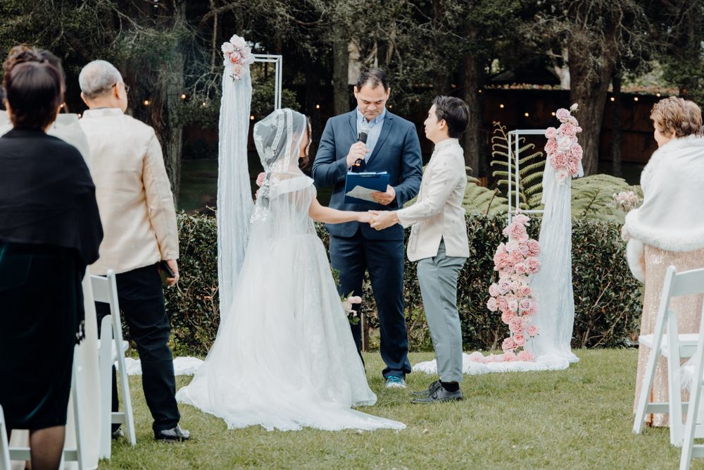 An intimate outdoor wedding ceremony takes place under trees with white decorative elements.