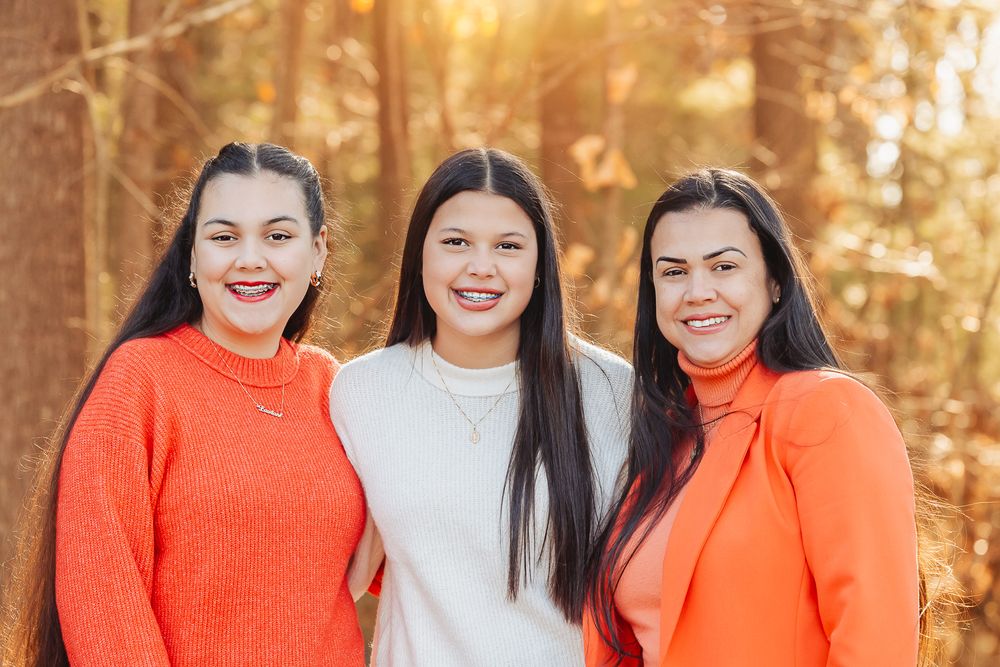 Three women in orange and white sweaters pose together in autumn woodland setting.