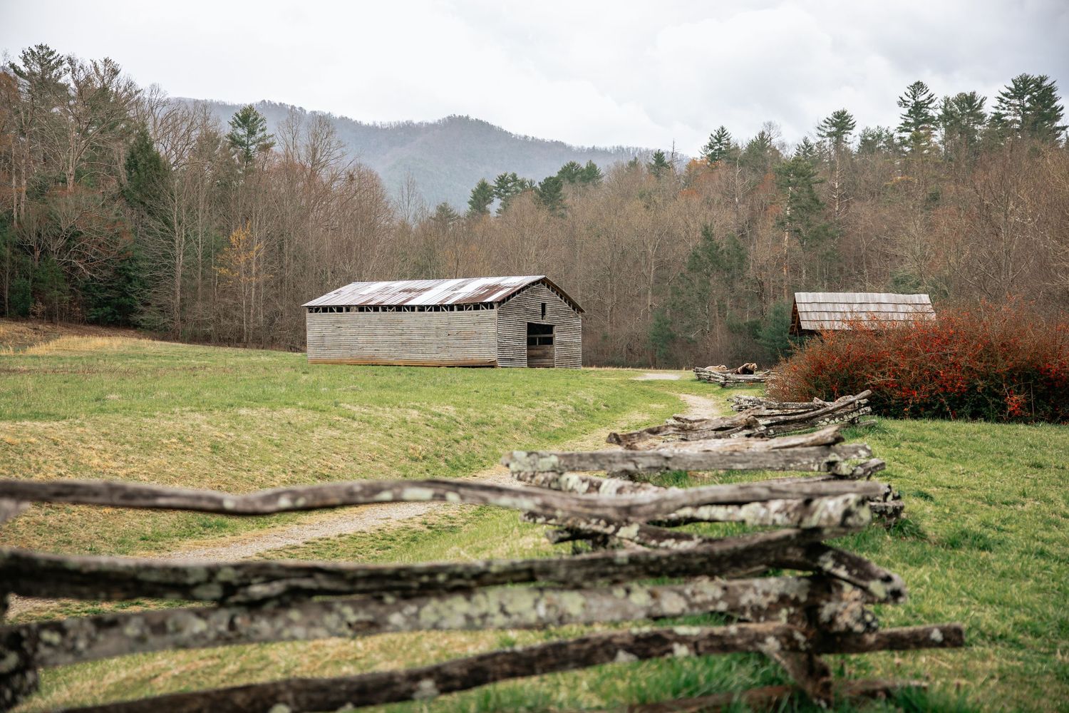 A rustic barn sits in a mountain valley with split-rail fence in the foreground and autumn trees in the background.