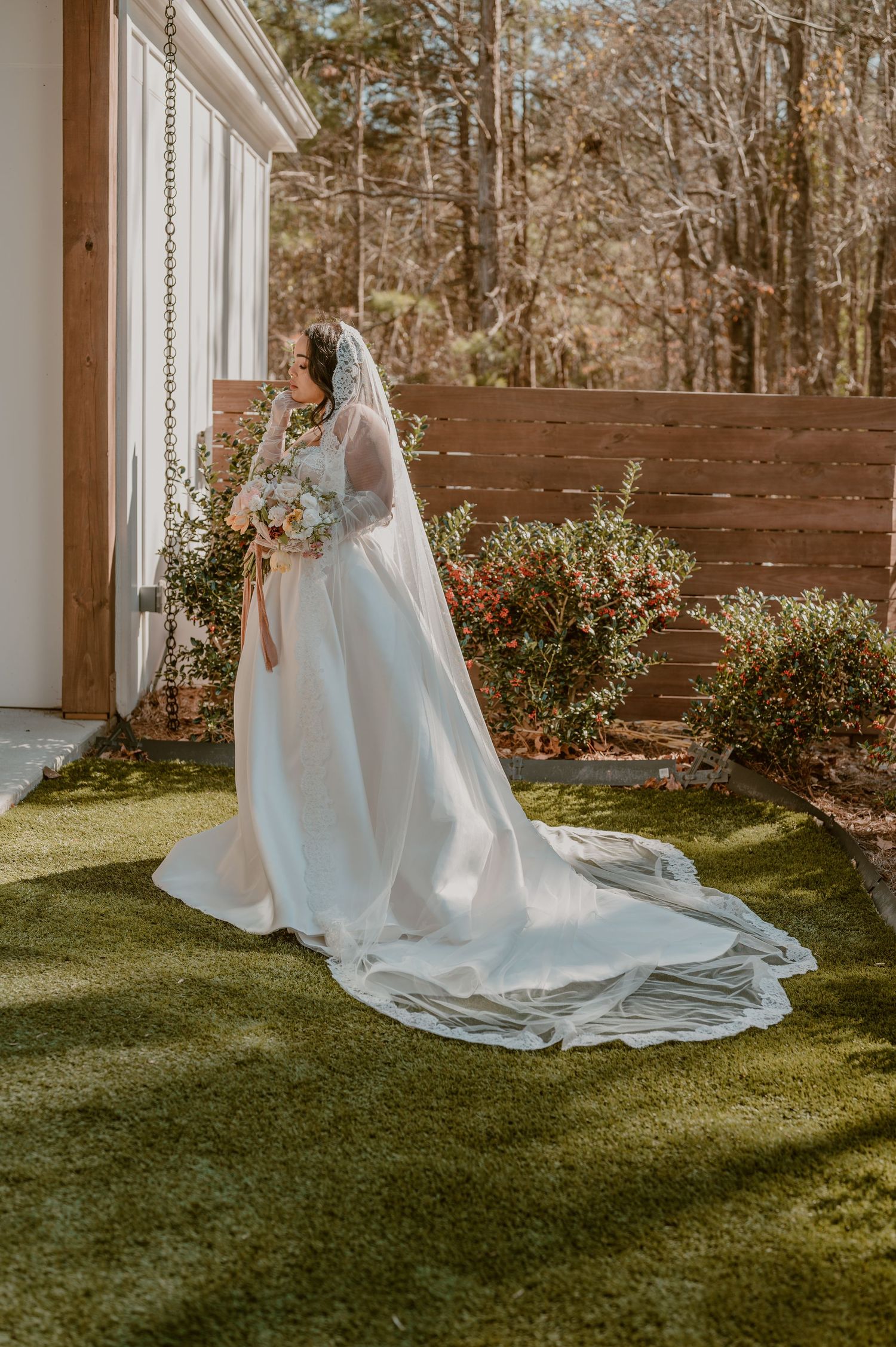 A bride in a flowing white wedding gown with a long veil and train stands in a sunlit garden with wooden fence backdrop.