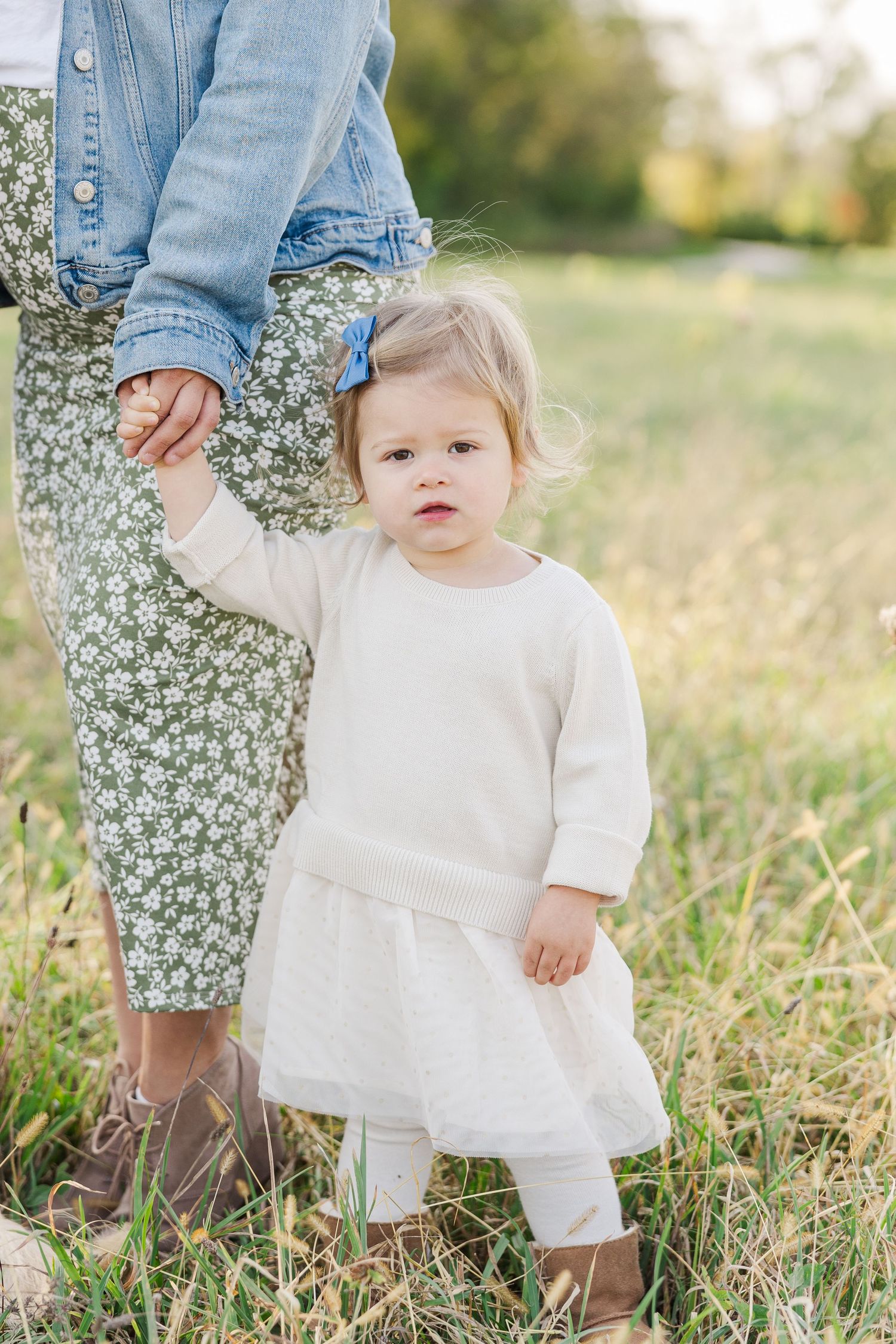 Small child in white dress and brown boots holds adult's hand while standing in a grassy field on sunny day.