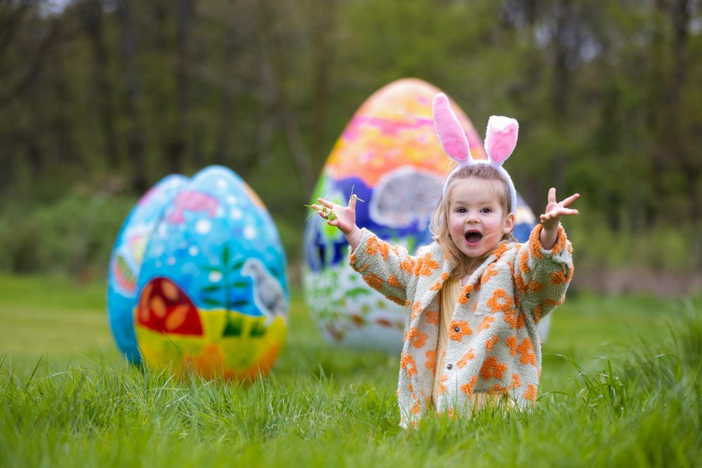 Child wearing bunny ears stands excitedly in a grassy field with large decorative Easter eggs in the background.