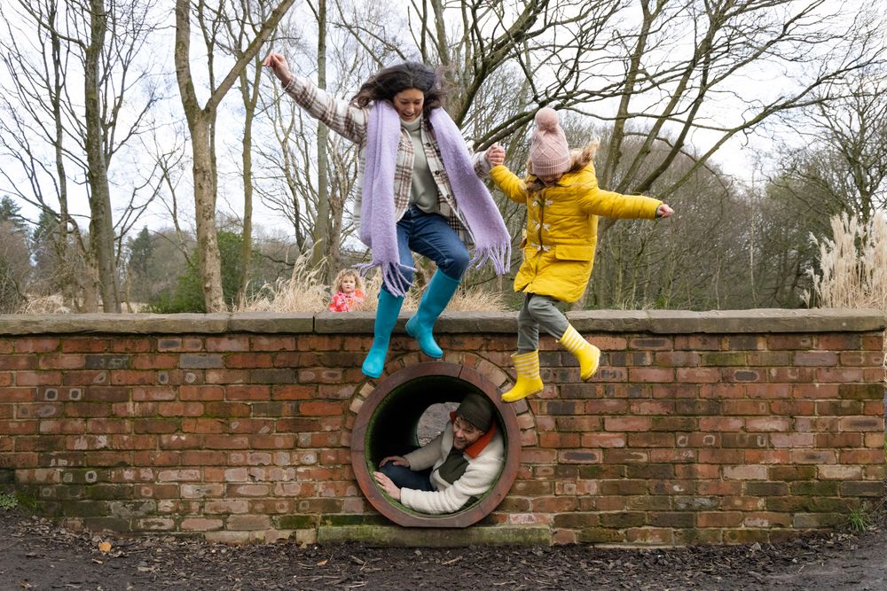 Two people in colorful rain boots and coats playfully pose on a brick wall with a circular drainage opening.