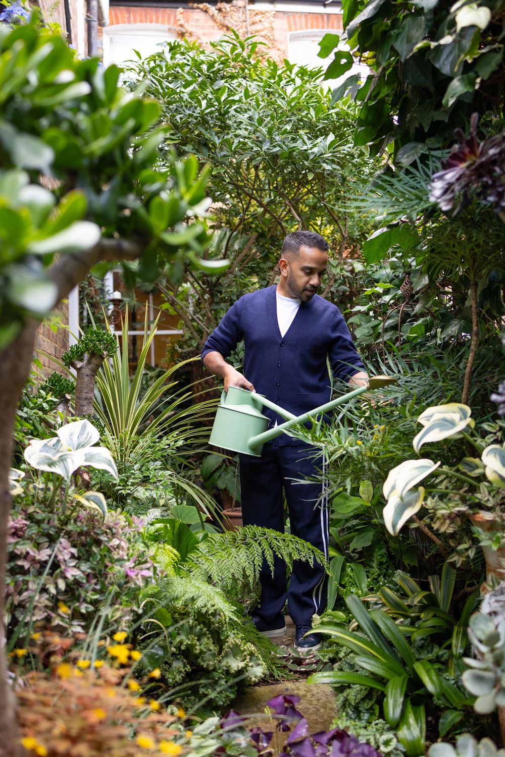 Someone tends to a lush garden filled with tropical plants and greenery while holding a watering can in a navy sweater.