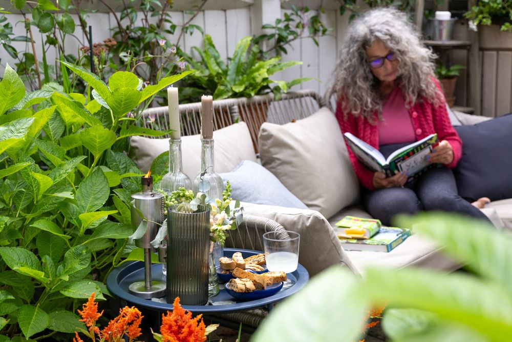 A tranquil garden patio scene with a cozy seating area, throw pillows, and vibrant plants surrounding a snack table.